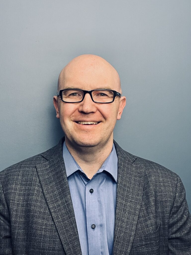 A smiling man wearing glasses and a blazer against a plain background.